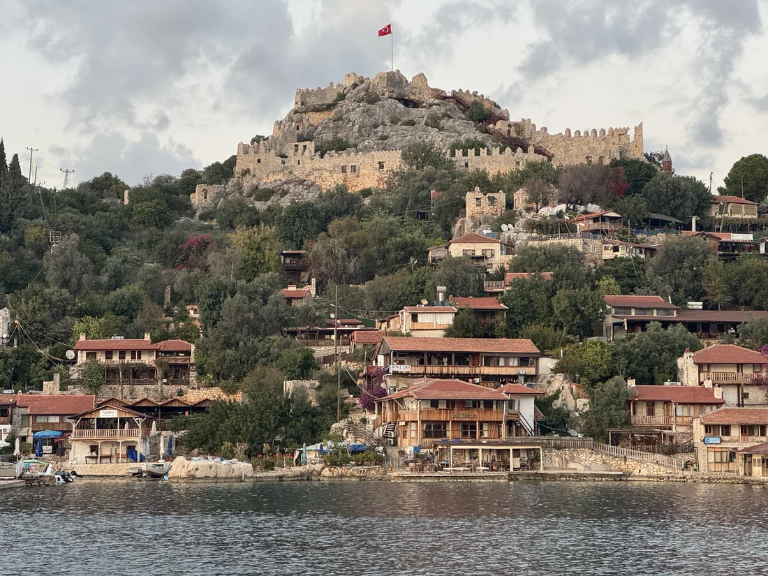 Häuser und Anlegestege von Kaleköy am Meer; auf dem Hügel darüber stehen die Burgmauern von Simena.